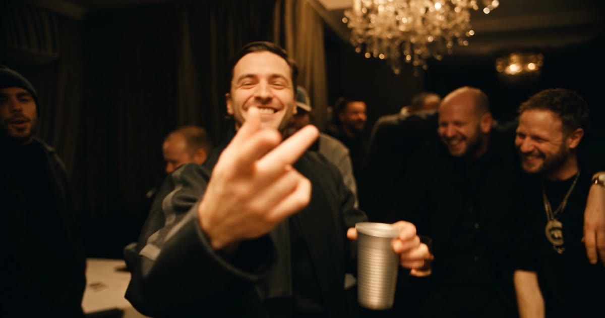 A Burning Bridges AI video: a man at the front of a party under crystal chandeliers, grinning and pointing at the camera with a silver cup in his hand while friends laugh behind him
