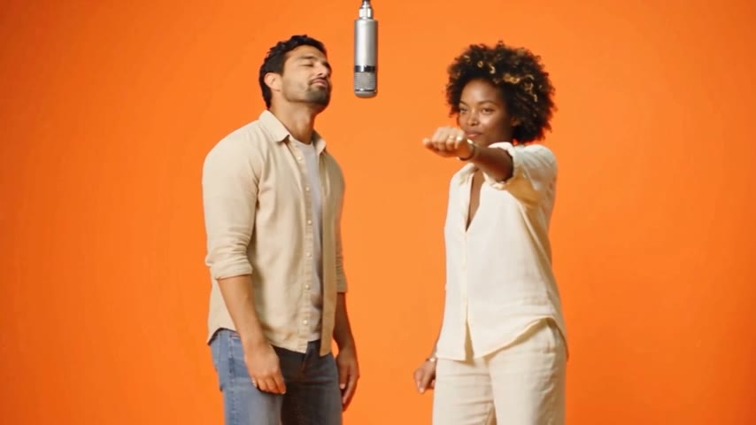 The same man and woman performing at a hanging microphone in front of an orange backdrop in the Hotel Lobby AI video